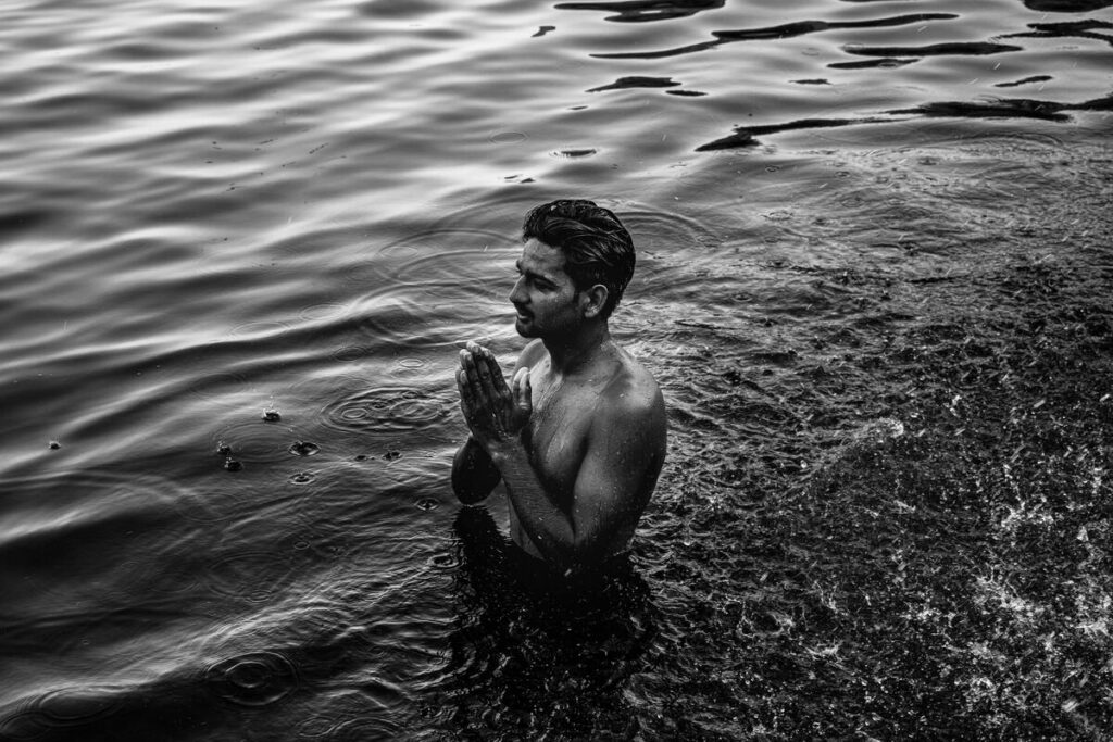 Homme debout dans l’eau, mains jointes en prière, regard vers l’horizon.