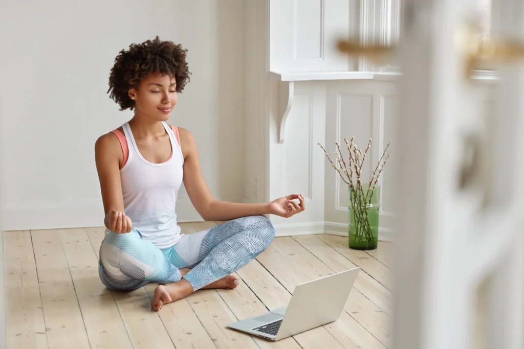 Femme assise en tailleur méditant devant un ordinateur portable pendant un cours de yoga en ligne.