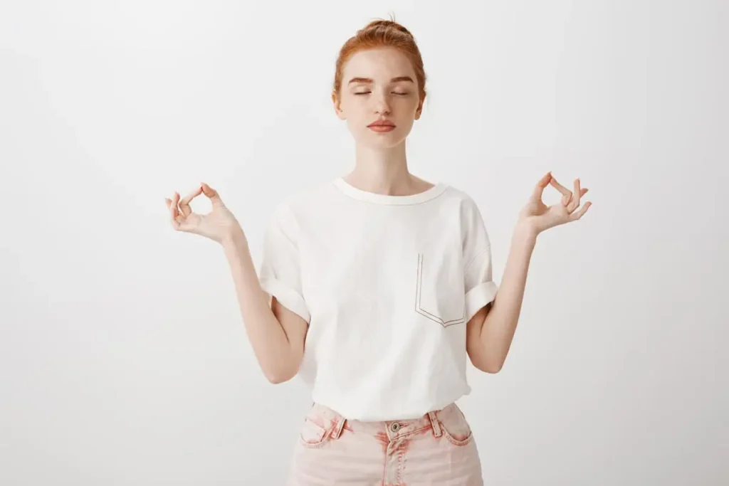 Femme rousse aux yeux fermés en posture de méditation debout, les mains en mudra, sur fond blanc