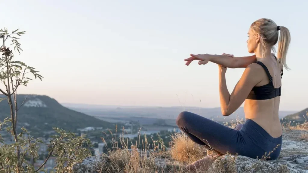 Femme blonde en tenue de sport pratiquant une posture de yoga assise en extérieur, face à un paysage naturel au lever du soleil