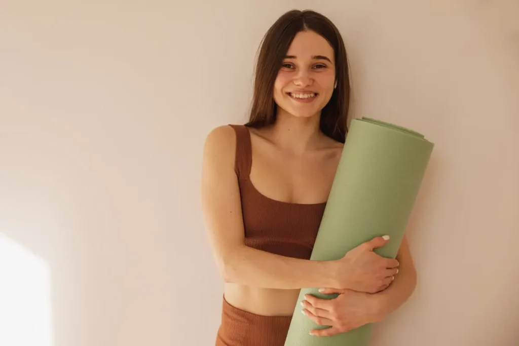 Jeune femme souriante en tenue de yoga tenant un tapis vert enroulé contre un mur beige